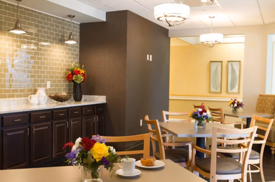 A dining area in Governor's Village featuring several tables with wooden chairs. Each table has a small floral centerpiece. The room has warm lighting with ceiling fixtures, a dark brown cabinet with a white countertop, and a tiled backsplash. The walls are painted in neutral tones with framed artwork visible in the background.