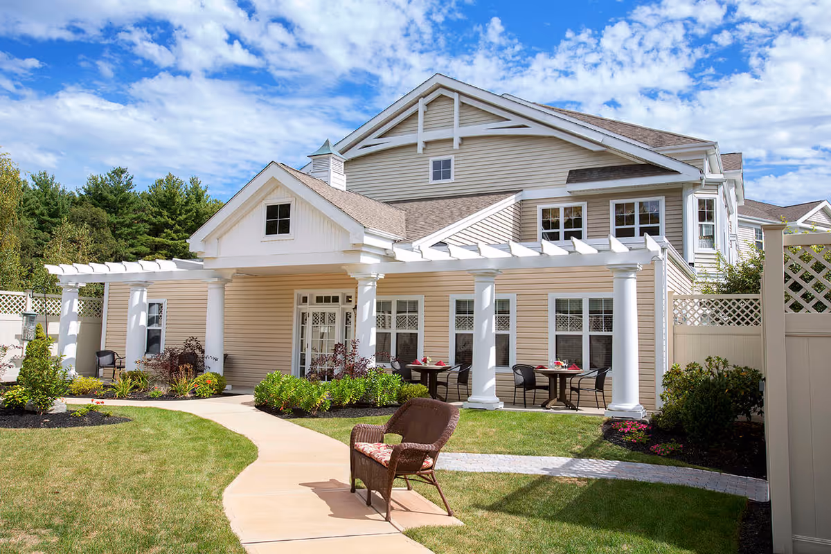 Exterior view of a senior living facility building with beige siding, white columns, and a pergola over the entrance. There is a curved walkway leading to the entrance, surrounded by green grass, shrubs, and small flower beds. Outdoor tables and chairs are set up near the windows under the pergola. The sky is partly cloudy with blue patches.