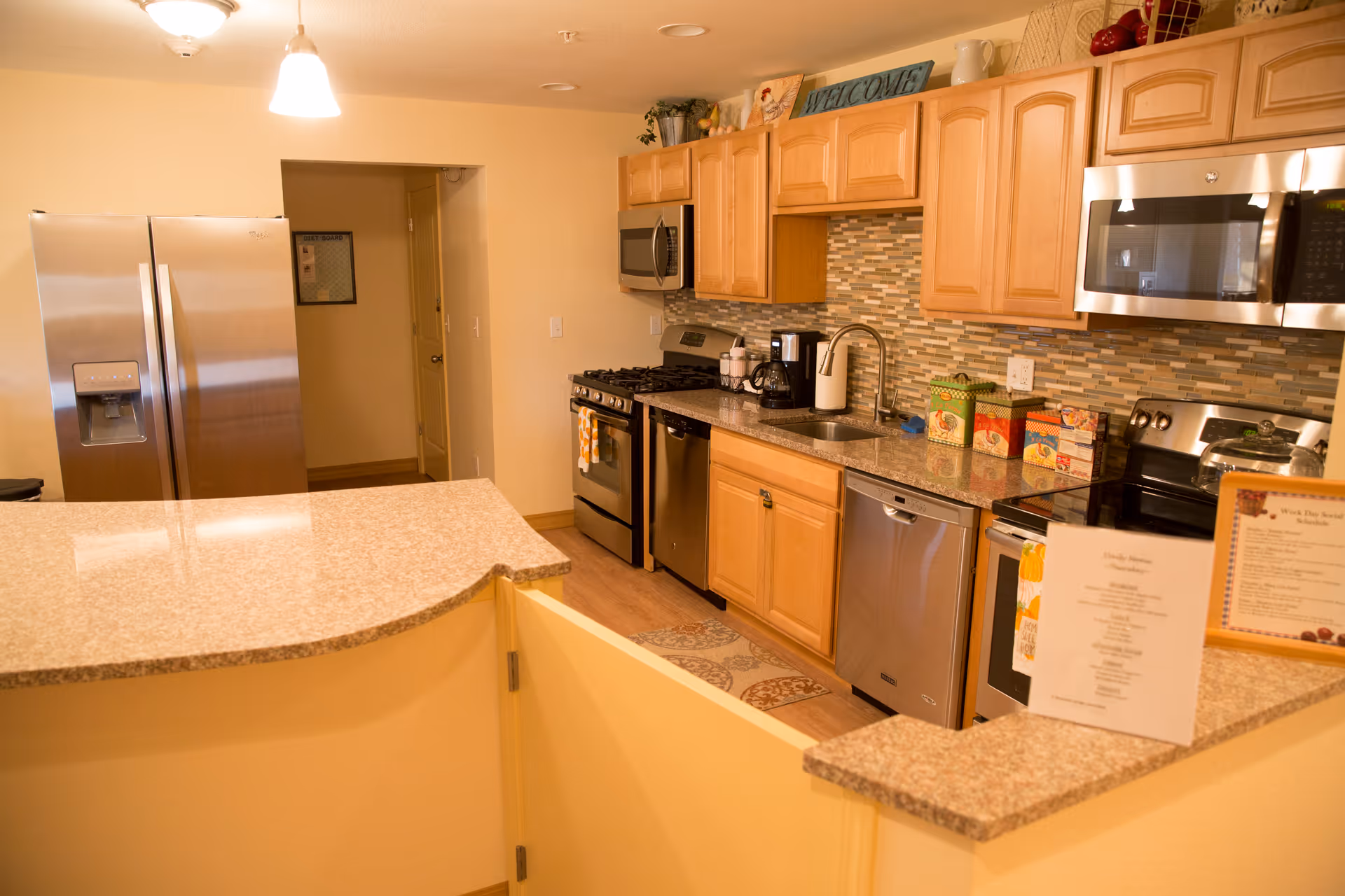 A modern kitchen with light wood cabinets, granite countertops, and stainless steel appliances including a refrigerator, stove, microwave, and dishwasher. There is a tiled backsplash and a small welcome sign on top of the cabinets. The kitchen is well-lit with ceiling lights and has a pass-through counter with a partial yellow safety gate.