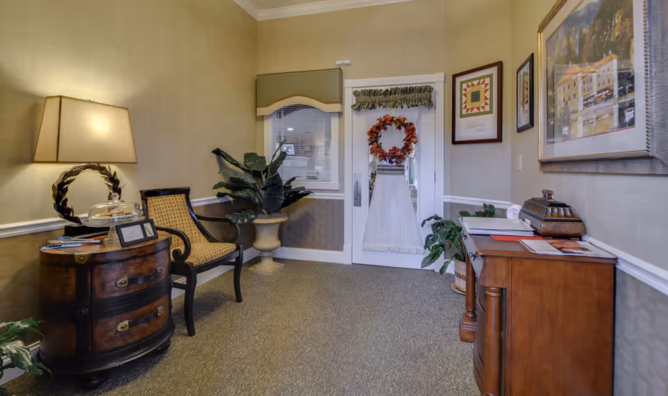 A cozy interior hallway or waiting area with beige walls and carpeted floor. There is a wooden side table with a lamp and a glass-covered plate, a cushioned chair with a woven back, a large potted plant, and a wooden cabinet with papers and a decorative box on top. A door with a window and a fall-themed wreath is visible at the end of the hallway, along with framed artwork on the walls.
