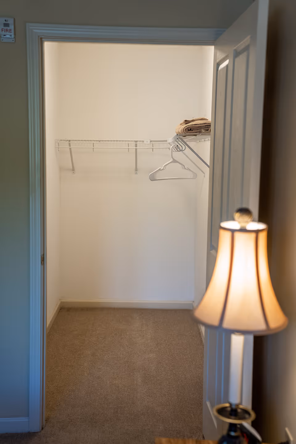 View of an empty walk-in closet with beige carpet, white walls, a wire shelf with a few hangers and folded towels, and a partially open door with a lit table lamp visible in the foreground.