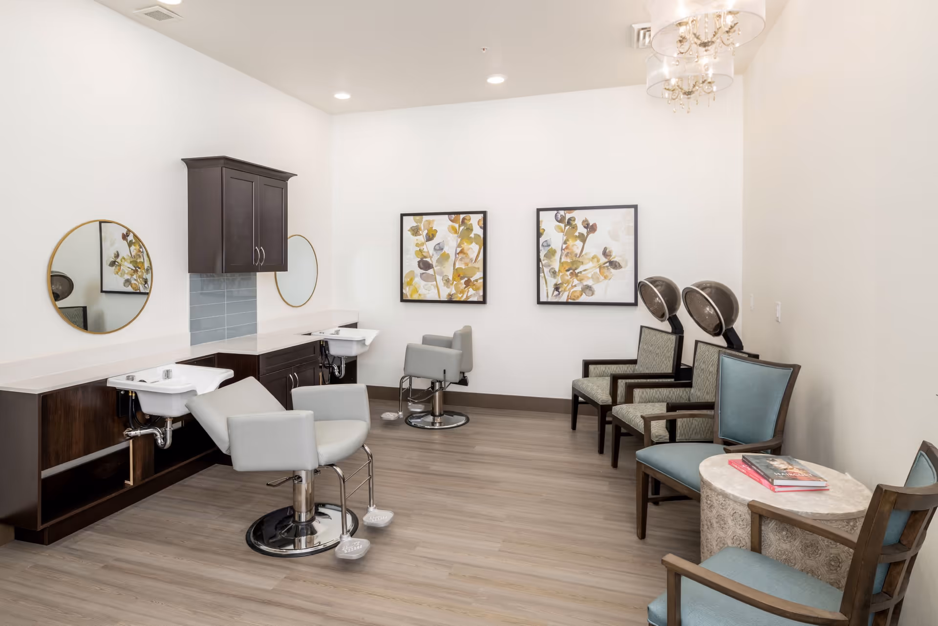 Interior view of a senior living facility's hair salon area with two salon chairs in front of sinks and mirrors, two hair drying chairs, and additional seating with a small round table holding books. The walls are white with two framed botanical prints and a chandelier hanging from the ceiling.