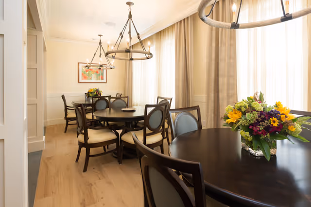 A bright dining room with multiple round dark wood tables and chairs with light-colored cushions. Large windows with sheer curtains allow natural light to fill the room. Two circular chandeliers hang from the ceiling, and a colorful flower arrangement is placed on the nearest table. A framed artwork is visible on the far wall.