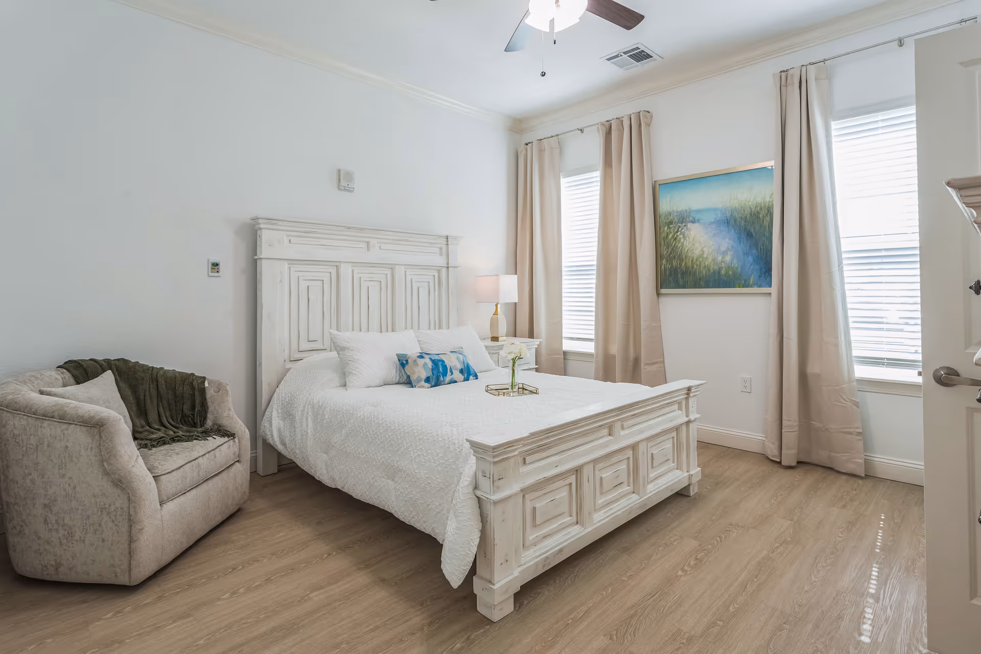 A bright bedroom with a white wooden bed frame and white bedding, accented with blue and white pillows. There is a beige armchair with a dark green throw blanket on the left side. Two windows with beige curtains and white blinds let in natural light. A landscape painting with green grass and a blue sky hangs on the wall between the windows. The floor is light wood, and a ceiling fan with a light is visible above.