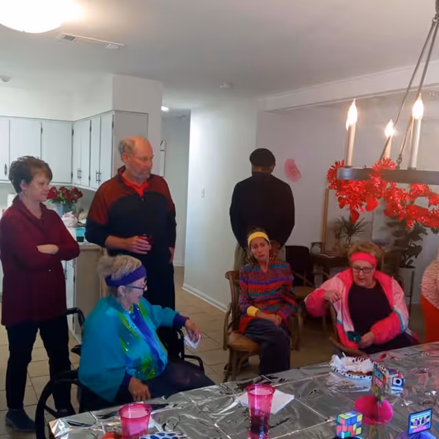 A group of elderly people gathered in a dining area with a table covered in a silver tablecloth. Some are seated while others stand, engaging in conversation. The room has white walls, a chandelier with red decorations, and a kitchen area visible in the background.