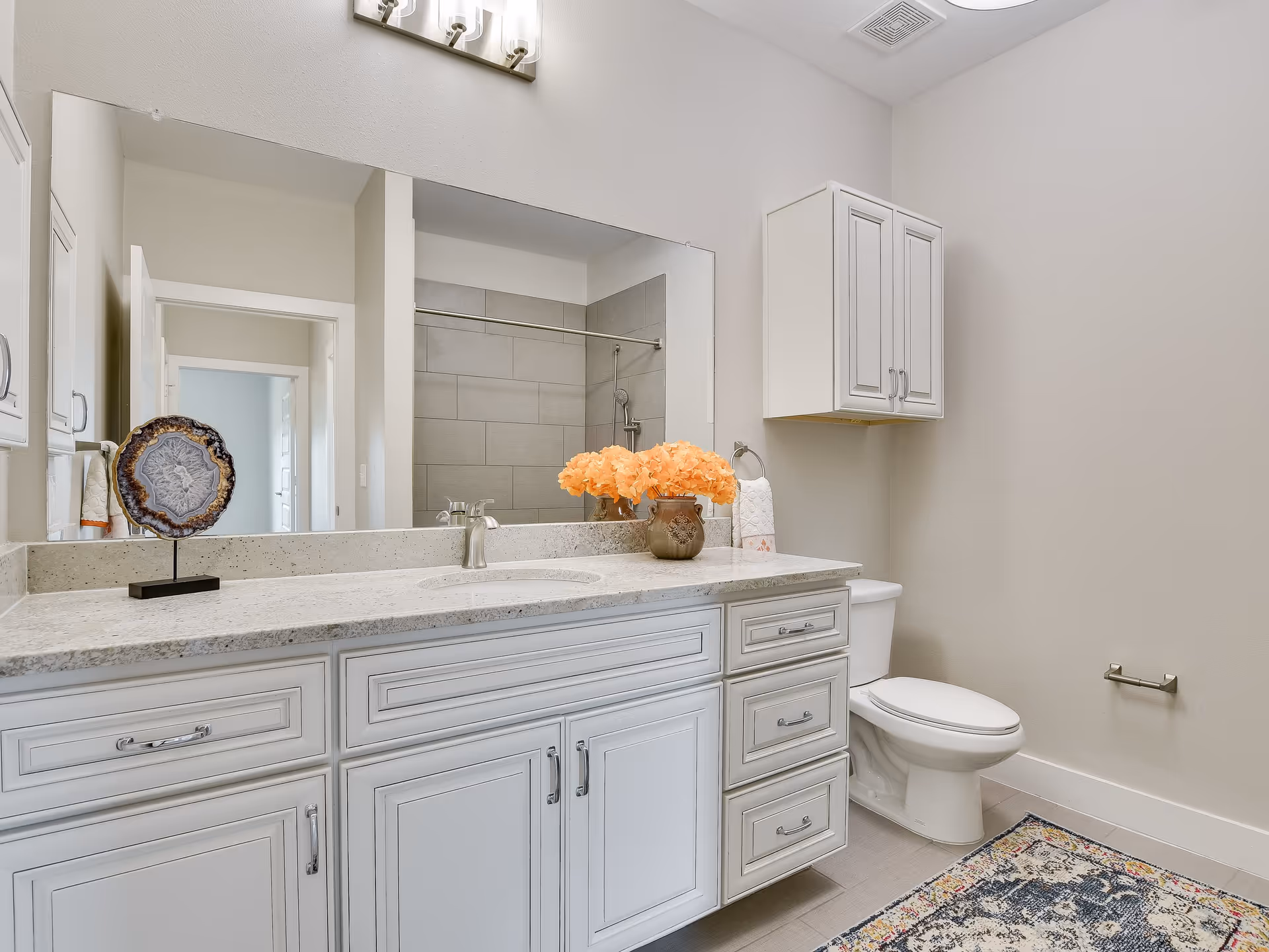 A clean and modern bathroom featuring a white vanity with multiple drawers and a granite countertop. On the countertop, there is a decorative vase with orange flowers and a decorative stone piece. Above the vanity is a large mirror with a light fixture mounted on the wall. To the right, there is a white toilet with a wall-mounted cabinet above it and a colorful patterned rug on the floor. The shower area with gray tiles and a showerhead is visible in the mirror's reflection.