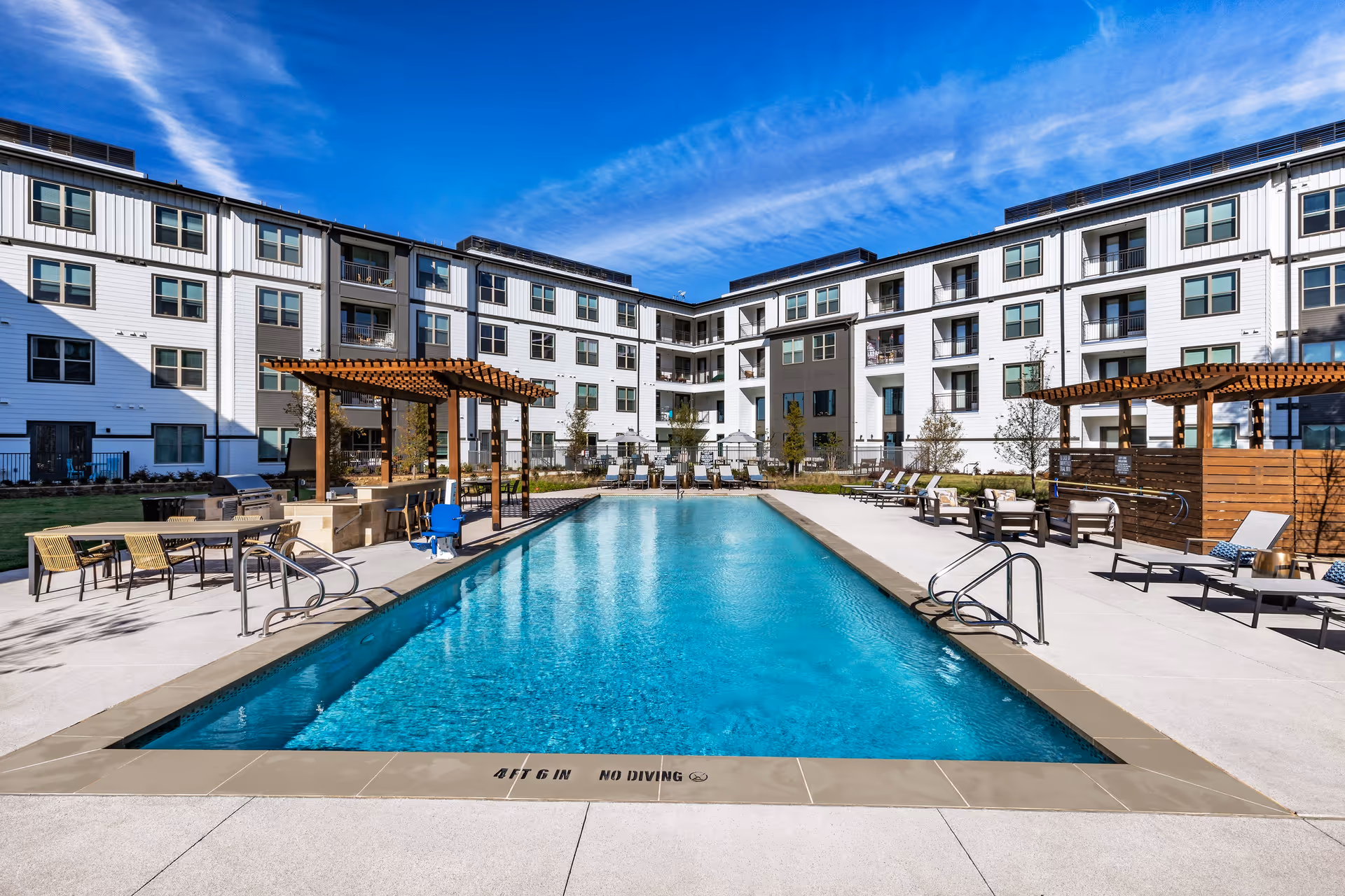 Rectangular outdoor swimming pool surrounded by lounge chairs and pergolas in the courtyard of a four-story residential building under a blue sky.