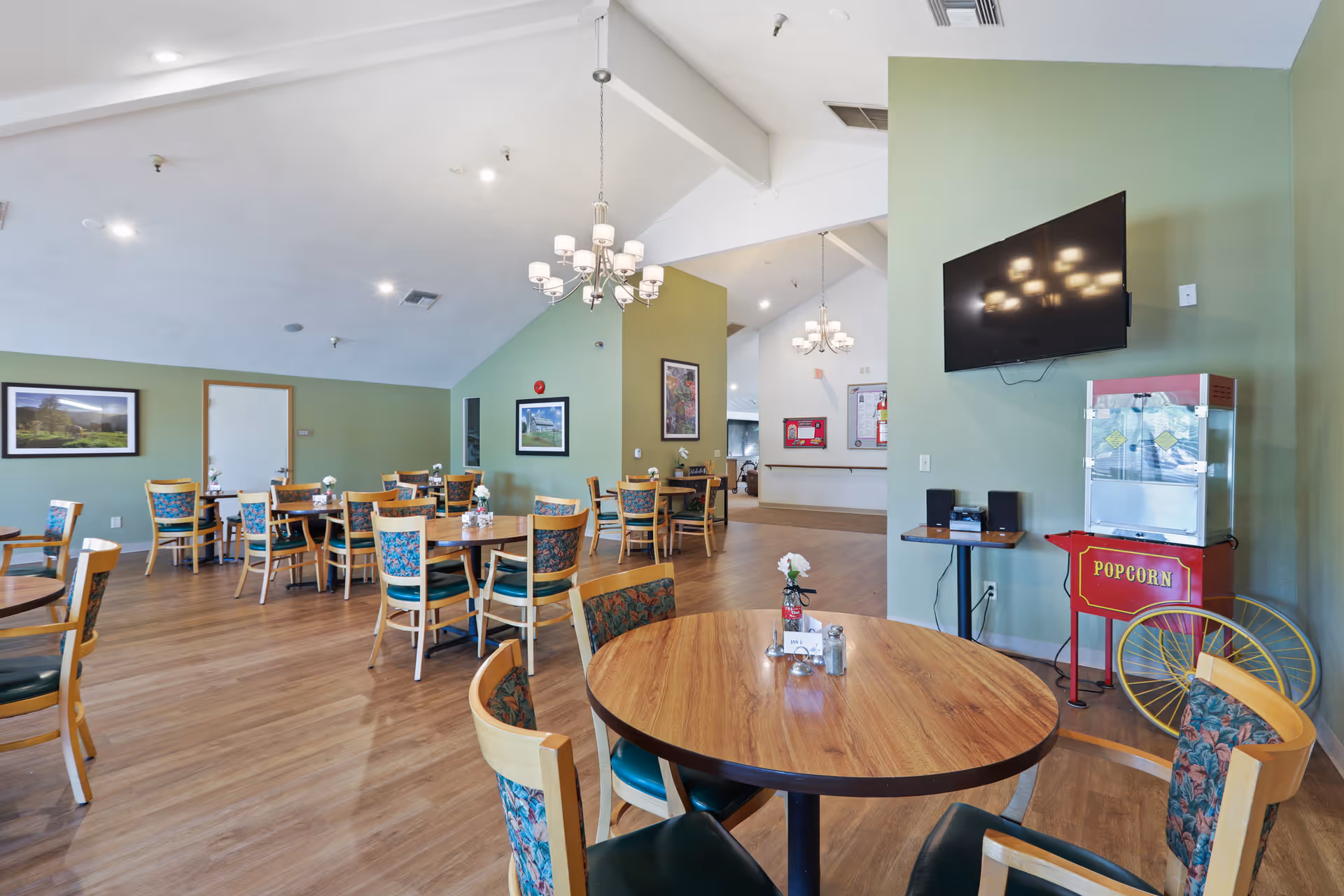 A spacious dining room with multiple round wooden tables and chairs with floral upholstery. The walls are painted green and decorated with framed pictures. There are two chandeliers hanging from the ceiling. A popcorn machine and a wall-mounted TV are visible on the right side of the room.