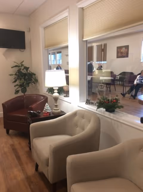 A cozy seating area in a retirement village with two beige armchairs and one brown leather armchair arranged around a small wooden table with a lamp and holiday decorations. A large window looks into another room where several people are seated. A flat-screen TV is mounted on the wall above a potted plant.