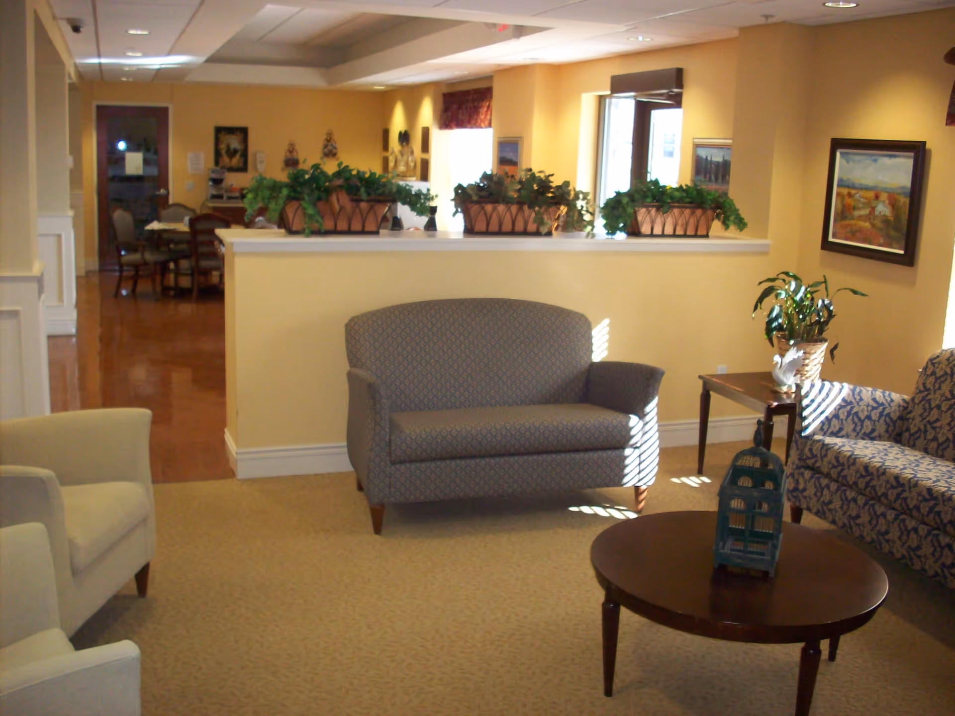 A cozy senior living facility common area with beige walls and carpet. The room features a patterned loveseat, a matching sofa, two beige armchairs, a wooden coffee table with a decorative birdcage, and a side table with a potted plant. There are several potted plants on a half wall divider, framed paintings on the walls, and a dining area visible in the background.