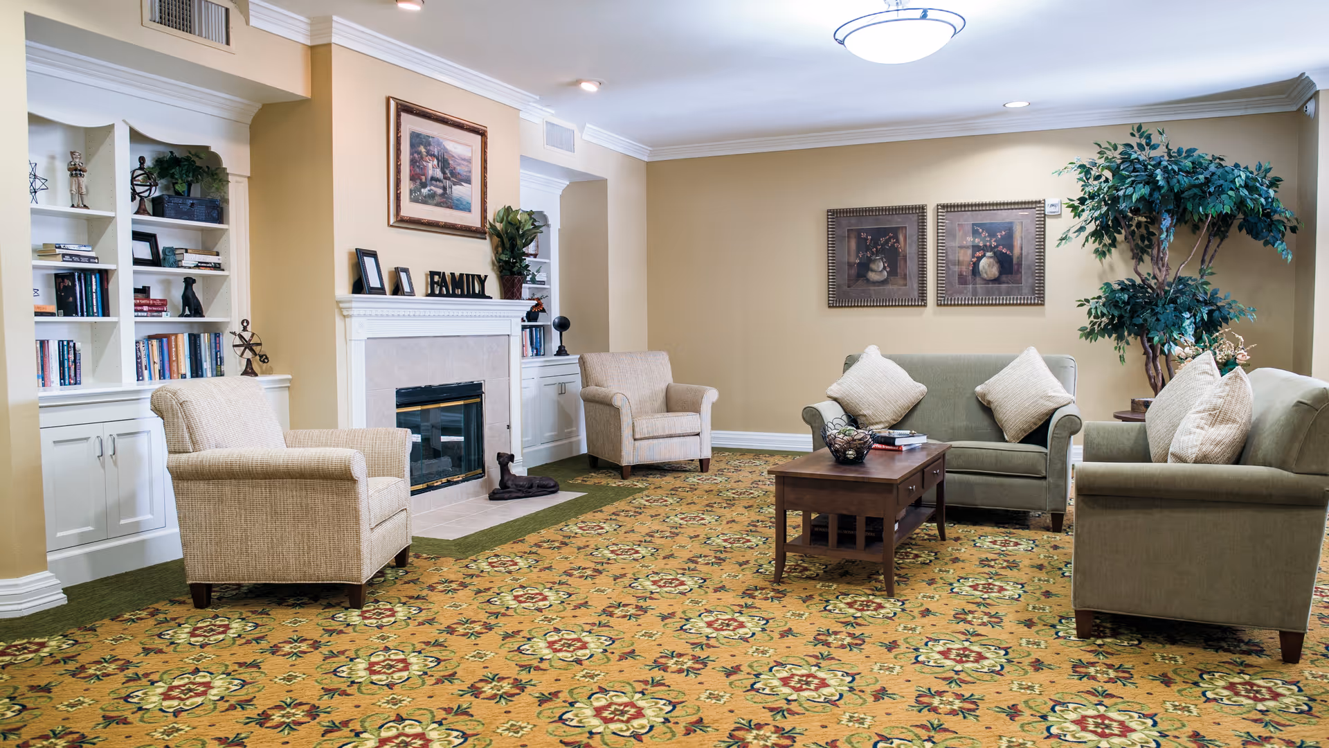 A cozy living room with beige walls and a patterned carpet. The room features two beige armchairs, two green sofas with beige cushions, a wooden coffee table with books and a decorative bowl, a fireplace with a mantle displaying picture frames and a 'FAMILY' sign, built-in white bookshelves filled with books and decorative items, framed artwork on the walls, and a large potted plant in the corner.