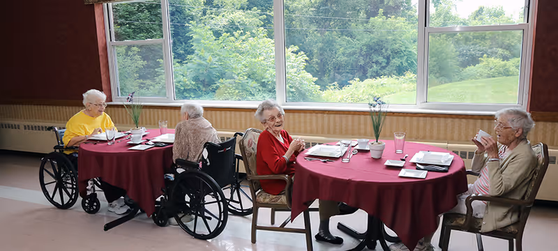 Four elderly women sitting around two round tables with red tablecloths in a dining area. Two women are in wheelchairs, and the other two are seated in chairs. The tables have plates, glasses, and small flower pots. Large windows behind them show green trees and a grassy area outside.