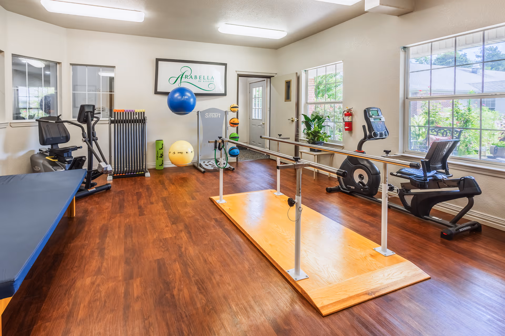A well-lit exercise room with wooden flooring featuring parallel bars on a wooden platform in the center, exercise bikes, a therapy table, various exercise balls, and fitness equipment. Large windows allow natural light to enter, and a sign on the wall reads 'Arabella of Kilgore'.