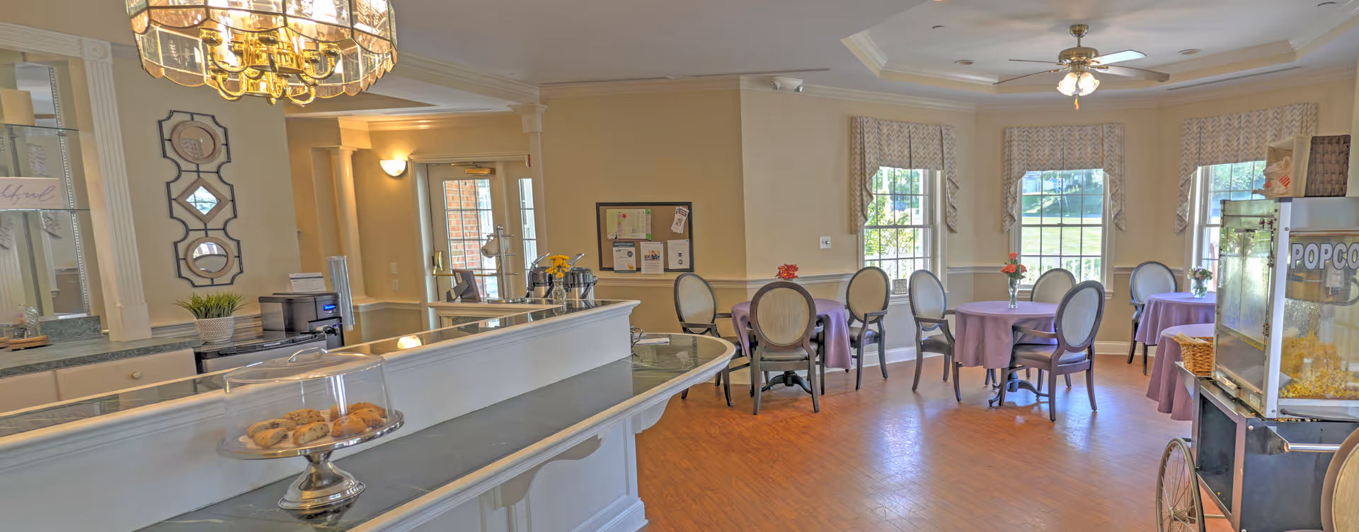 A bright dining area with round tables covered in purple tablecloths and surrounded by chairs. There are windows with patterned valances letting in natural light. A popcorn machine is visible on the right side, and a counter with a glass dome containing cookies is in the foreground. The room has hardwood floors, a ceiling fan, and a chandelier above the counter.