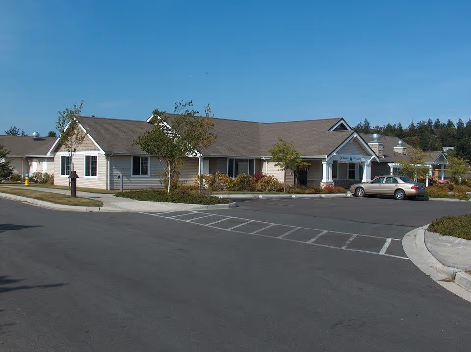 Exterior view of a single-story building with beige siding and a brown roof, surrounded by a parking lot with a few cars and some landscaping including small trees and bushes under a clear blue sky.