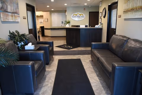 Reception area of Hope Stay Memory Care facility with a dark wood front desk, a black leather sofa and two black leather armchairs, a black rug on the floor, potted plants, and framed artwork on the walls.