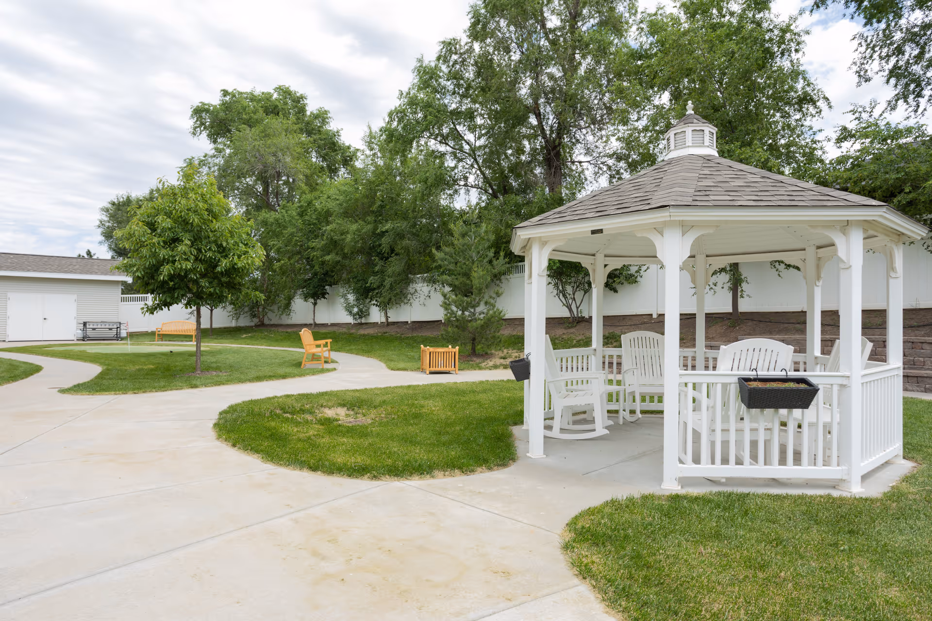 Outdoor garden area with a white gazebo containing white rocking chairs and a table, surrounded by green grass, trees, and a paved walkway. There are additional benches and a small shed in the background under a cloudy sky.
