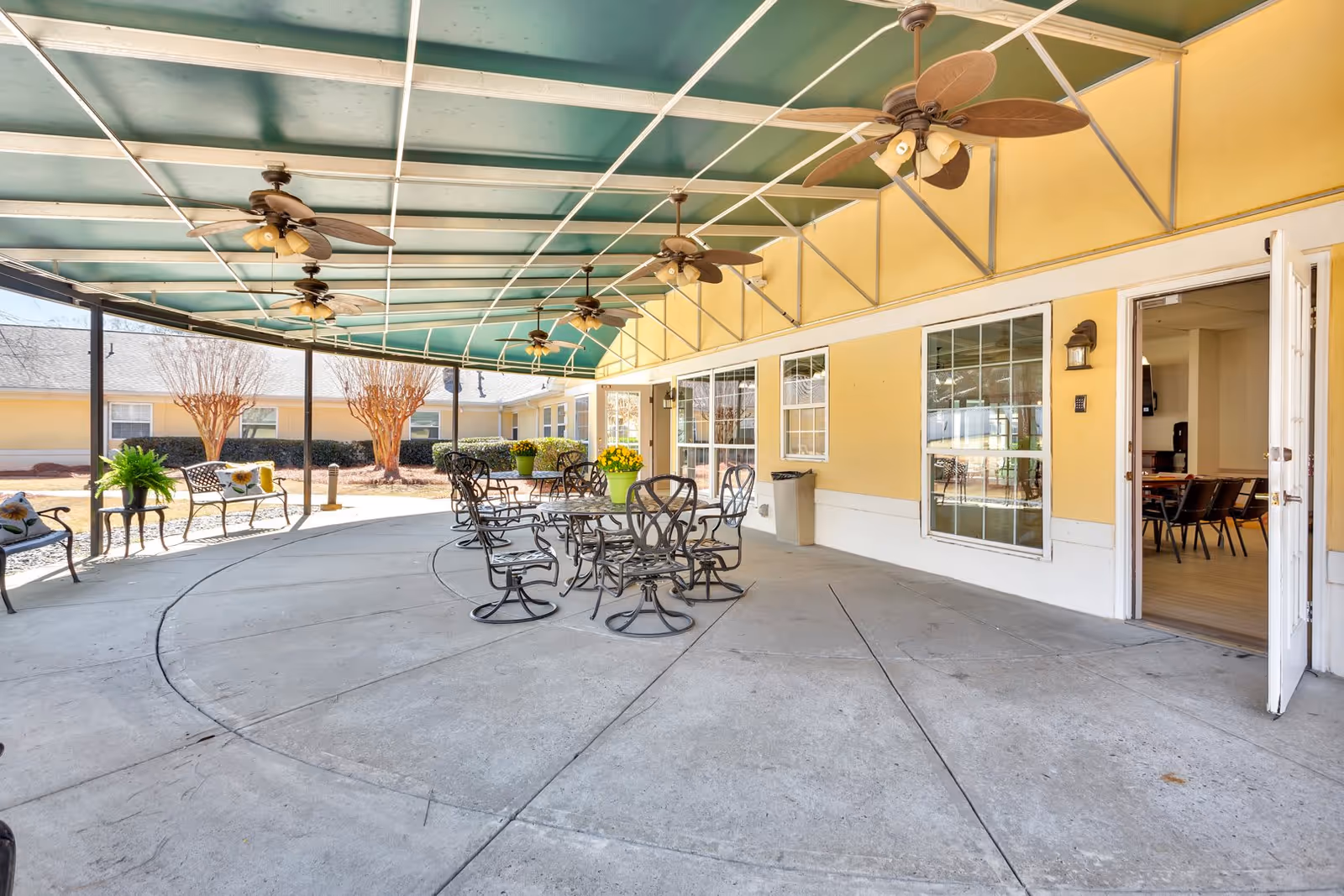 Covered outdoor patio area with several ceiling fans, metal tables and chairs, and benches with cushions. The patio is adjacent to a yellow building with large windows and an open door leading inside. There are potted plants on the tables and some landscaping with bushes and trees in the background.