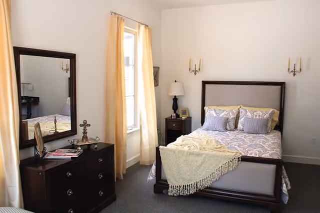 A bright bedroom with a large bed featuring patterned bedding and multiple pillows. There is a wooden dresser with a mirror on the left side, decorated with a cross and some magazines. Light yellow curtains frame a window letting in natural light. A nightstand with a lamp and picture frame is beside the bed. The walls are white and the floor is carpeted in gray.