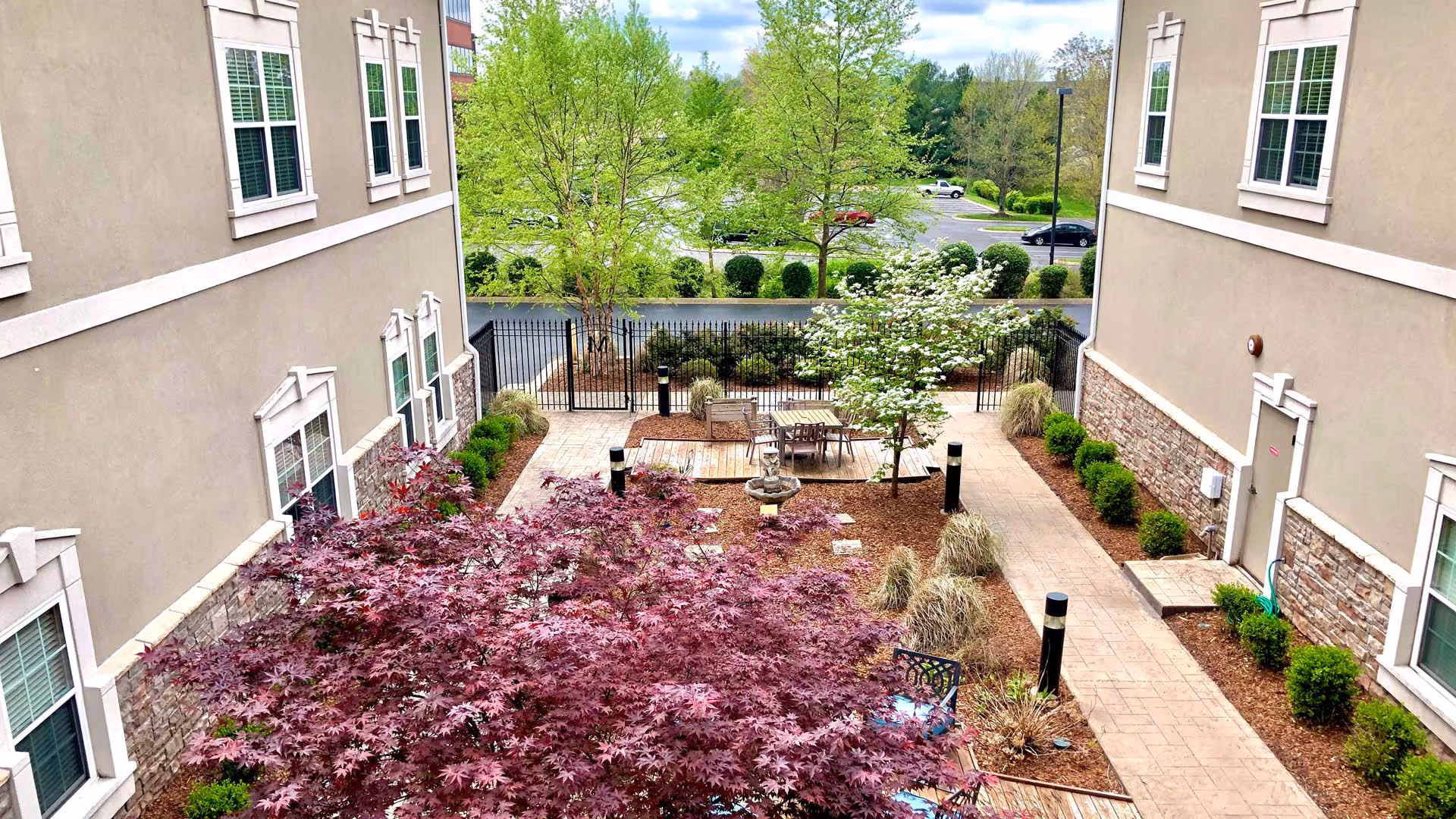 Outdoor courtyard area between two beige buildings with stone accents, featuring a paved walkway, landscaped garden beds with bushes and small trees, a seating area with a table and chairs, and a black metal fence in the background with a parking lot and green trees beyond.