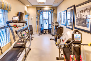 A small fitness room with exercise equipment including a treadmill, stationary bike, elliptical machine, and a multi-functional weight machine. The room has beige walls, two windows with blinds, and framed black and white photographs on the right wall.
