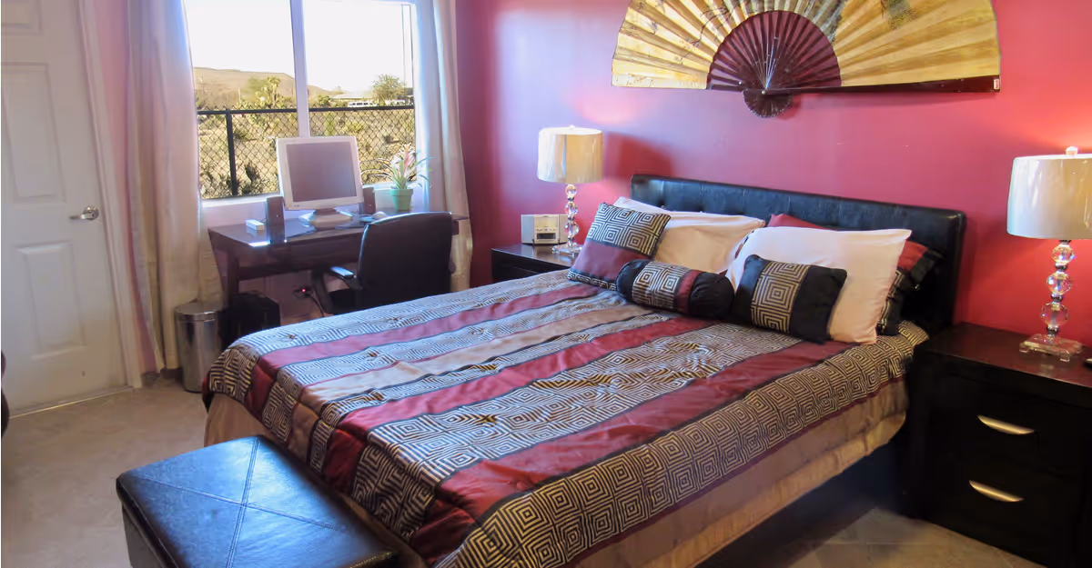 Bedroom with a patterned bed and pillows against a red accent wall, two lamps on nightstands, a desk by the window, and a decorative fan above the headboard.
