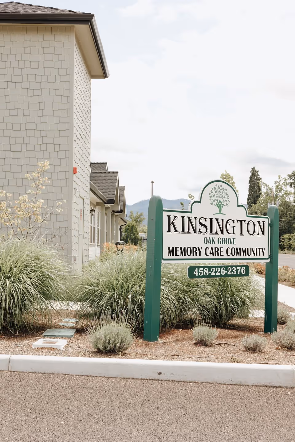 A green-and-white sign reading "Kinsington Oak Grove Memory Care Community" stands amid landscaping in front of a light-gray building.