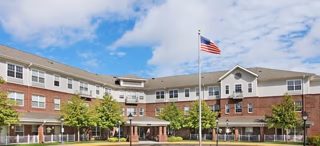 Front exterior view of a three-story senior living facility building with a brick and white siding facade, several windows, a covered entrance, an American flag on a flagpole, green trees, and a well-maintained lawn under a partly cloudy blue sky.