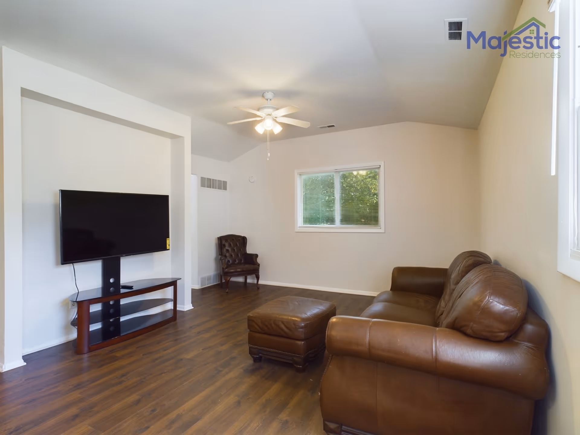 A living room with a brown leather sofa and matching ottoman, a dark brown armchair in the corner, a flat-screen TV mounted on a stand against a white wall, wood flooring, a ceiling fan with lights, and a window with blinds showing greenery outside.