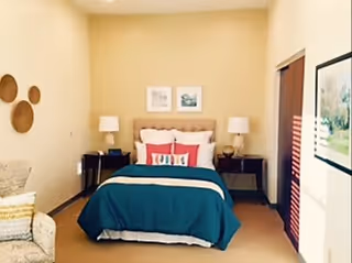 A neatly made bed with teal bedding and coral pillows centered between two nightstands and lamps in a warmly lit bedroom.