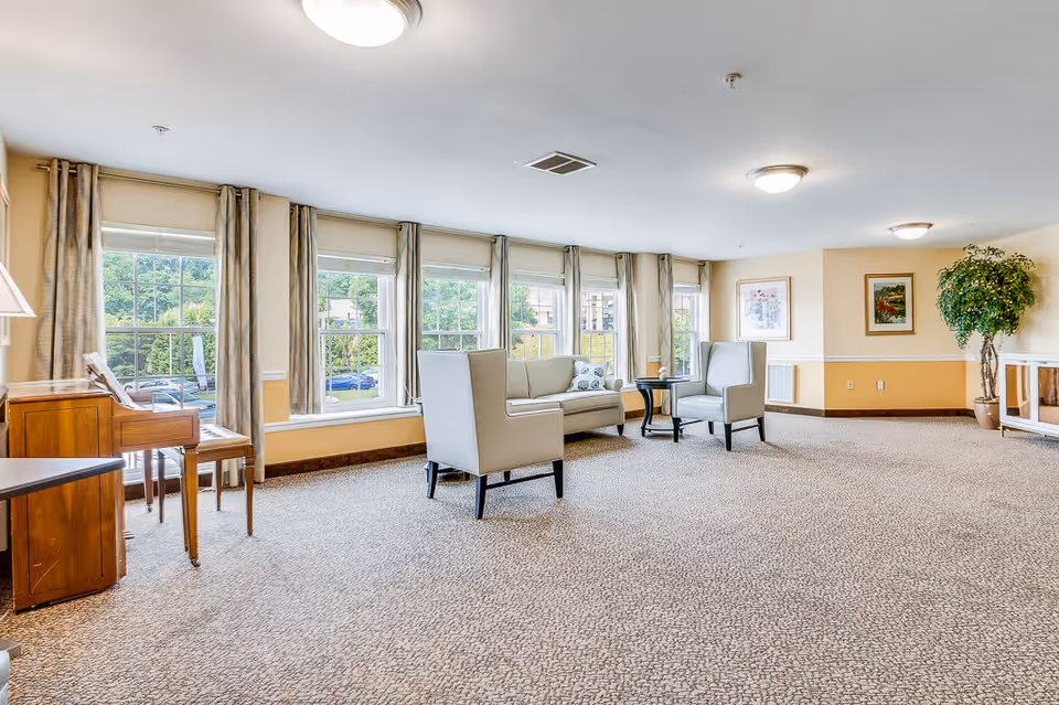 A spacious, well-lit living room area with large windows covered by curtains allowing natural light to fill the room. The room features a beige carpet, two light gray armchairs, a matching sofa, a small round black table, a wooden piano with a bench, framed artwork on the walls, and a tall potted plant in the corner.