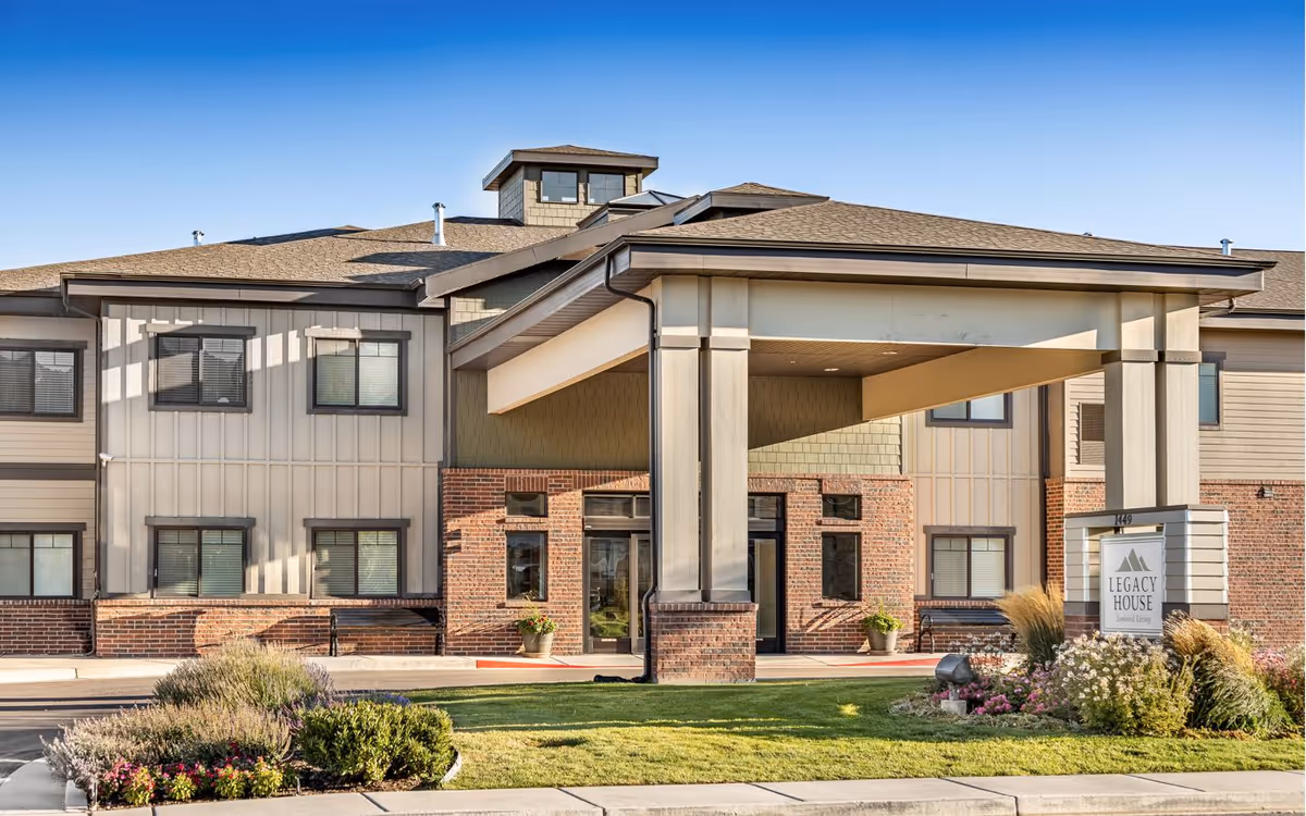 Exterior view of Legacy House of Spanish Fork, a senior living facility with a covered entrance, brick and siding facade, multiple windows, and landscaped greenery including bushes and flowers in front.