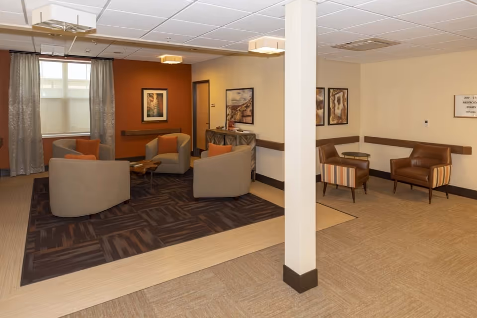 A cozy seating area in a senior living facility with four beige armchairs arranged around a wooden coffee table on a dark patterned rug. The walls are painted in warm tones with framed artwork, and there are two additional brown leather chairs with striped sides and a small round table in the corner. The space is well-lit with ceiling lights and has a window with curtains.