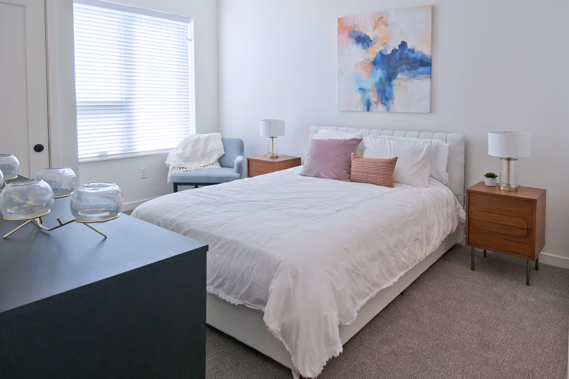 A bright and modern bedroom with a white bed featuring white and purple pillows, two wooden nightstands with lamps, a blue armchair with a white throw blanket, a large window with white blinds, a dark dresser with decorative glass candle holders, and an abstract painting with blue and pink tones on the wall.