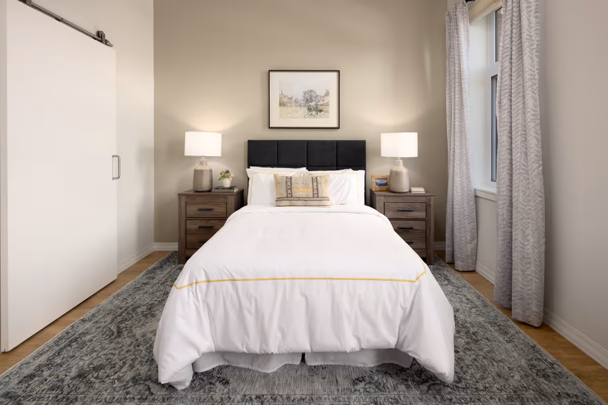 A neatly made bed with white bedding and a decorative pillow in a bedroom. The bed has a black upholstered headboard and is flanked by two wooden nightstands, each with a lamp. There is a framed artwork hanging on the beige wall above the bed. A window with patterned curtains is on the right side, and a sliding door is on the left. The floor has a patterned rug.