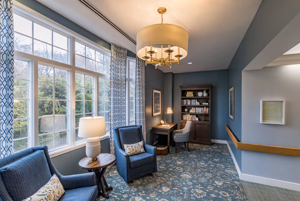 A cozy sitting area with two blue upholstered armchairs and a small round wooden table with a lamp between them. Large windows with patterned curtains let in natural light. In the background, there is a wooden bookshelf filled with books and decorative items, and a small desk with a chair and a desk lamp. The walls are painted blue, and the floor is covered with a patterned carpet. A modern chandelier hangs from the ceiling.