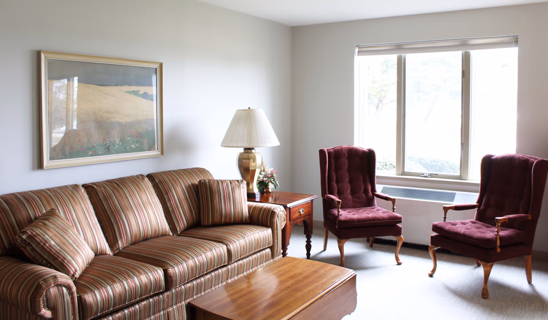 Sunlit living room with a striped sofa, wooden coffee table, side table with a lamp and flowers, and two burgundy armchairs by a window.