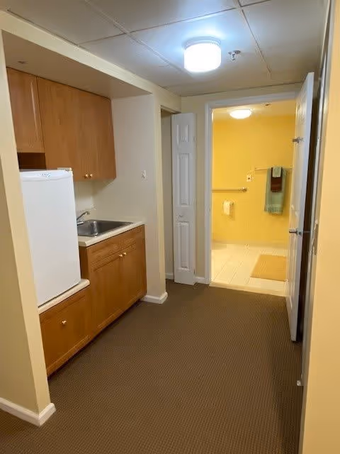 A small kitchenette area with wooden cabinets, a white mini refrigerator, and a sink. The kitchenette opens into a hallway leading to a bathroom with yellow walls, a towel rack with towels, and a grab bar near the toilet.