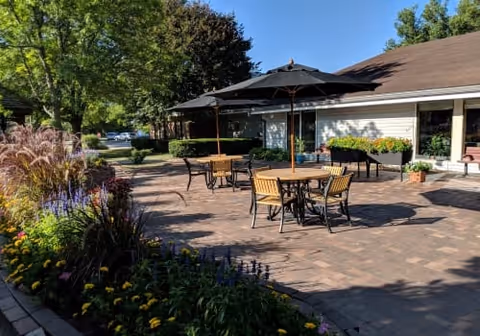 Outdoor patio area with several tables and chairs under large black umbrellas, surrounded by flower beds and greenery, adjacent to a single-story building with windows and a brown roof.