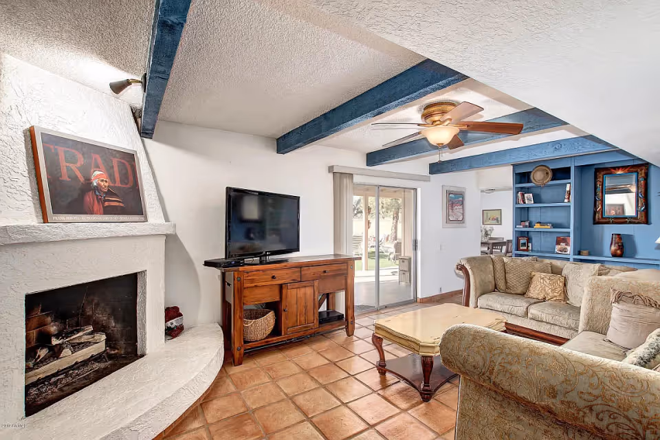 A cozy living room with terracotta tile flooring, a white textured fireplace with a framed artwork on the mantel, a flat-screen TV on a wooden console, a beige sectional sofa with patterned cushions, a wooden coffee table, blue built-in shelves with decorative items, and a ceiling fan with wooden blades. Sliding glass doors lead to an outdoor area.