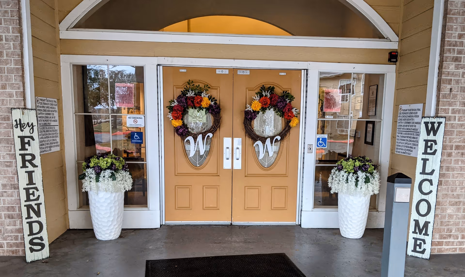 Entrance to a building with double orange doors decorated with floral wreaths featuring the letter W. On either side of the doors are large white planters with green and purple flowers. Two vertical signs flank the entrance, one reading 'Hey FRIENDS' and the other 'WELCOME'. Glass panels with handicap accessibility signs are on both sides of the doors.
