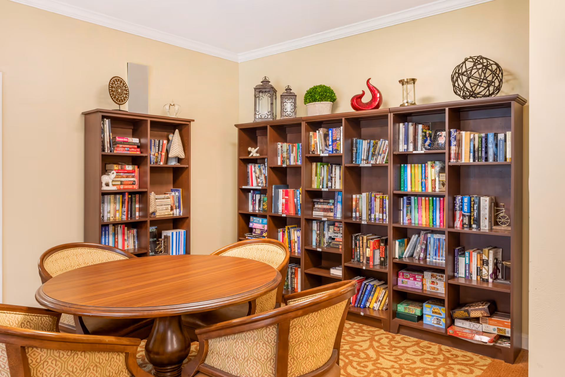 A cozy reading room with a round wooden table surrounded by four upholstered chairs. Behind the table are multiple dark wooden bookshelves filled with books, board games, and decorative items including lanterns, a green plant, and various sculptures. The room has beige walls and a patterned carpet.