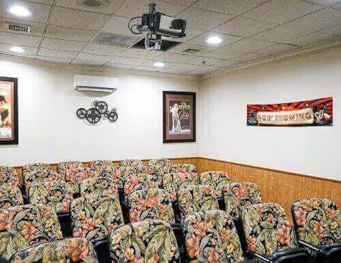 Small screening room with rows of floral-upholstered chairs, a ceiling-mounted projector, and framed posters on the walls.