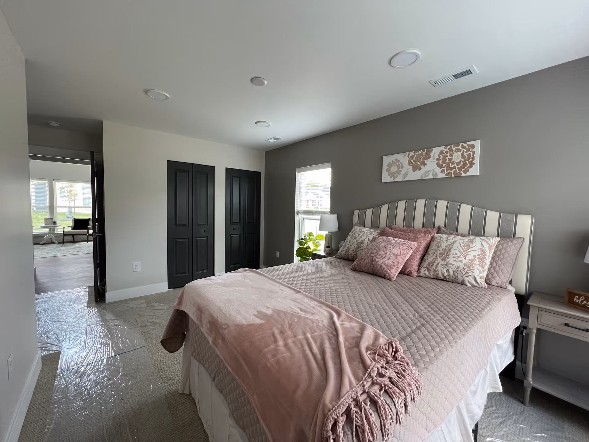 Well-lit bedroom with a striped headboard bed dressed in a pink quilt and floral pillows, a nightstand, window, and two dark closet doors.