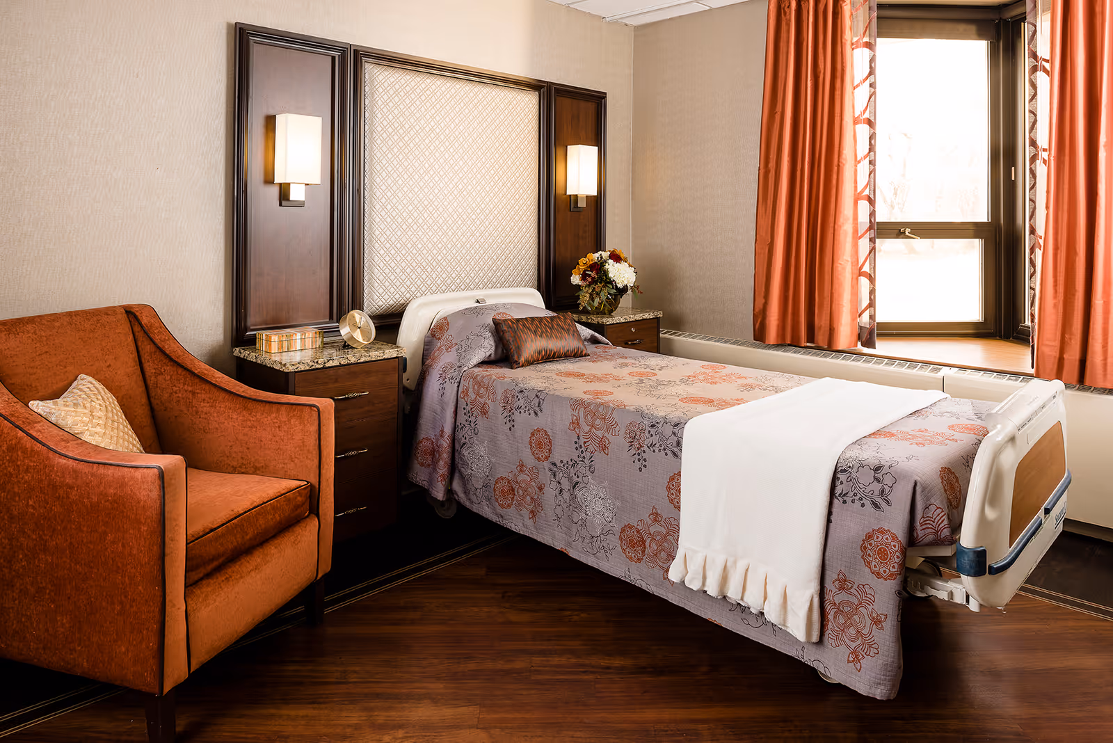 A cozy bedroom in a senior living facility featuring a single bed with a floral patterned bedspread and a white blanket at the foot. The bed is flanked by two wooden nightstands with granite tops, each with a wall-mounted light above. There is an orange upholstered armchair with a beige pillow to the left of the bed. A window with orange curtains allows natural light into the room, and a vase with flowers sits on one of the nightstands.