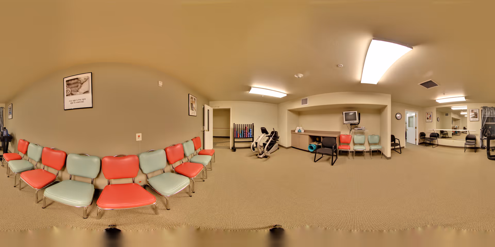 A spacious room with beige walls and carpeted floor, featuring a row of red and light blue chairs lined up against one wall. The room contains exercise equipment including a stationary bike and a rack of colorful weights. There is a small TV on a shelf, a large mirror on one wall, and fluorescent ceiling lights providing illumination. The room appears to be a fitness or activity area within a senior living facility.