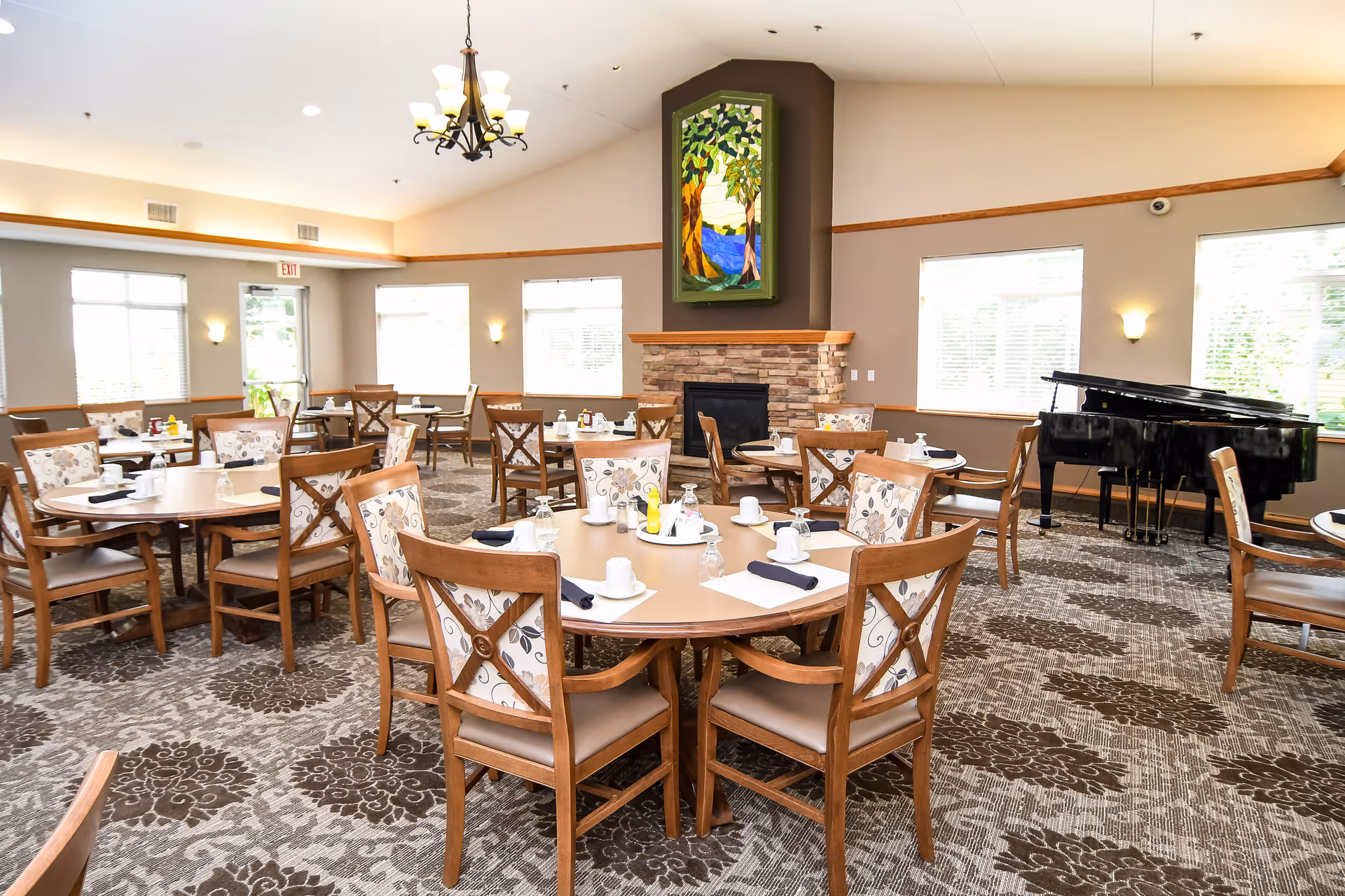 Bright dining room with round tables and chairs, a fireplace topped by stained-glass artwork, and a grand piano.