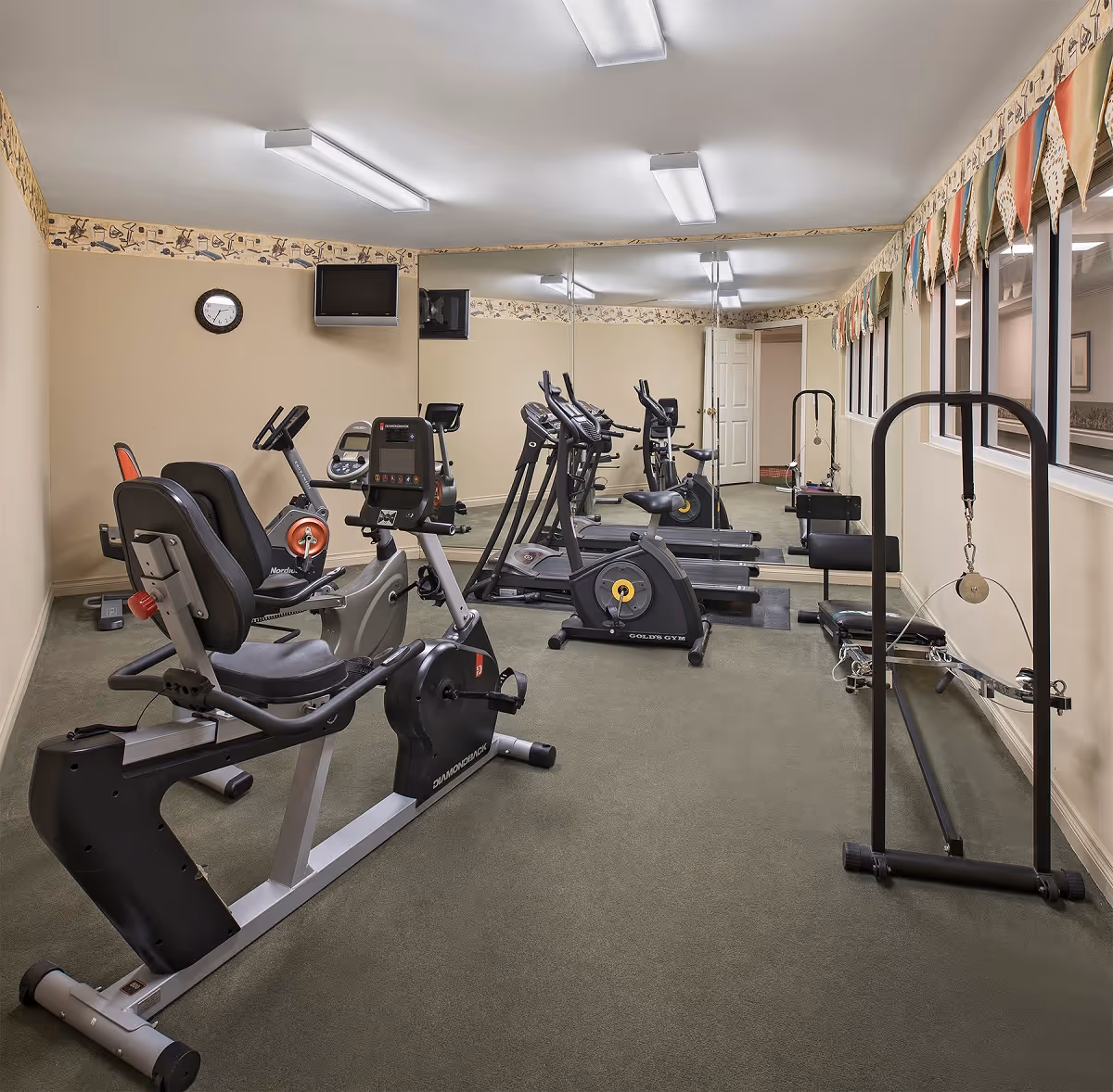 A small fitness room with exercise equipment including stationary bikes, elliptical machines, and a pulley system. The room has beige walls with a decorative border near the ceiling, a wall clock, a mounted TV, and large mirrors covering one wall. There are windows with colorful valances on the right side.