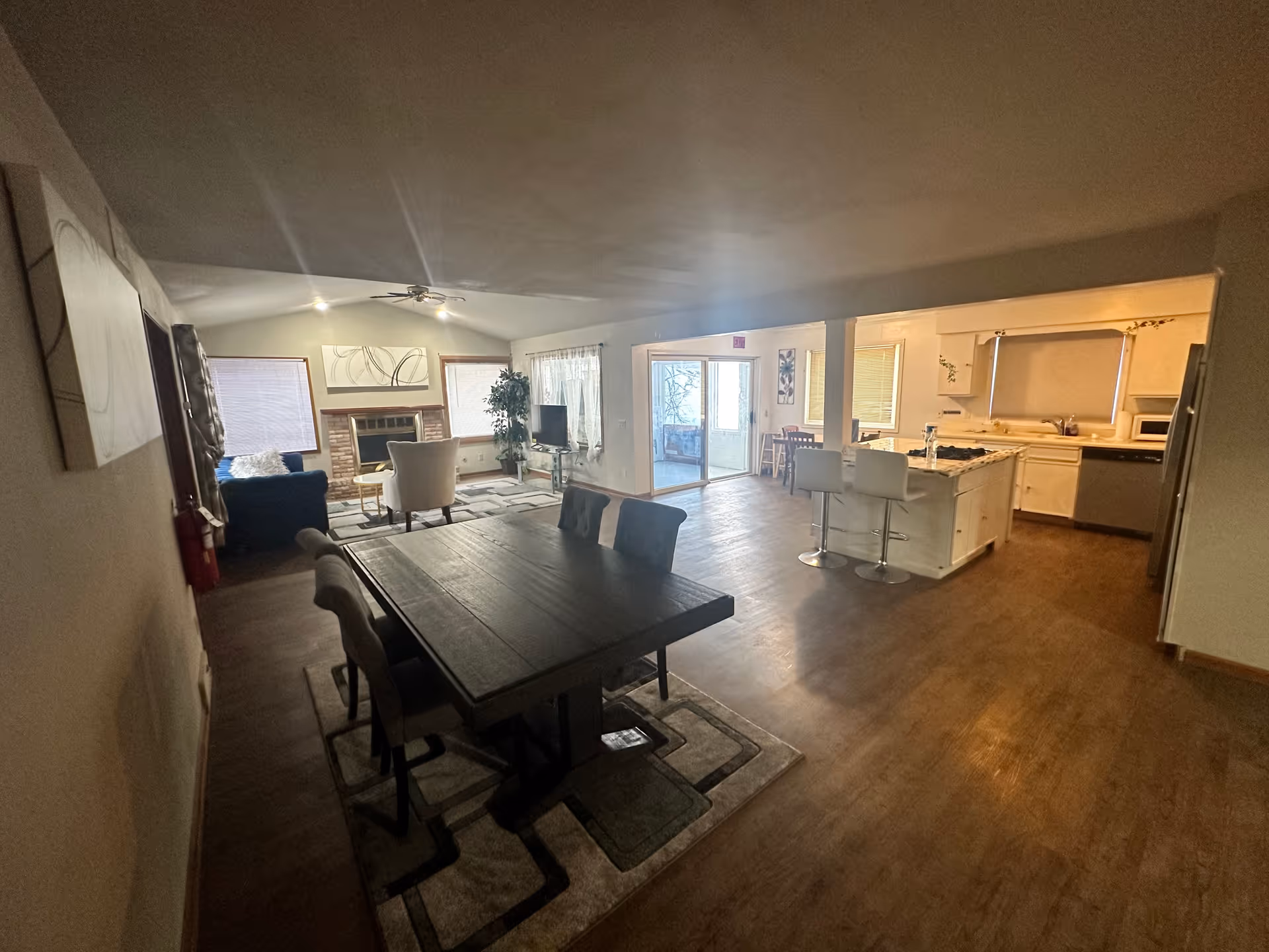 Open interior space showing a dining area with a dark wooden table and six chairs on a patterned rug, a living room with a blue sofa, white armchair, coffee table, TV on a stand, and a brick fireplace, and a kitchen with white cabinets, a kitchen island with two white bar stools, and stainless steel appliances. There are windows with blinds and a sliding glass door leading to another room.
