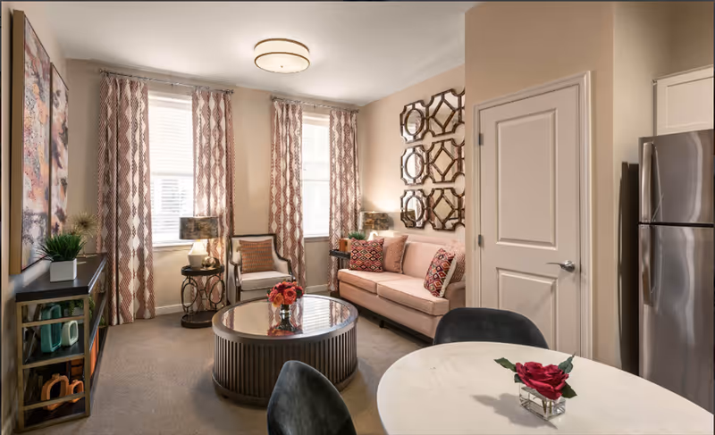 A cozy living room area with a beige sofa adorned with patterned cushions, a round glass coffee table with a flower vase, two armchairs, and a small round dining table with two chairs. The room has three windows with patterned curtains, a decorative wall mirror, a stainless steel refrigerator, and a door. The walls are painted in a light beige color.